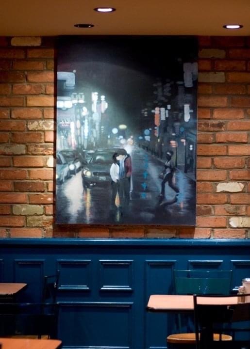 Susan's painting of a couple on a rainy, neon-lit city street, hanging on a brick wall at Williams Fresh Cafe